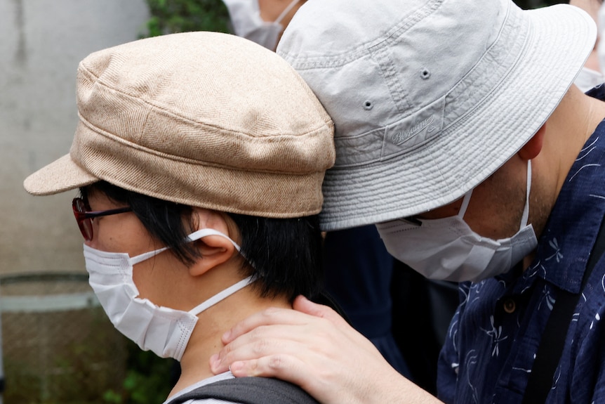 A masked Asian man bows his head and gently holds the shoulder of the capped and masked woman in front of him