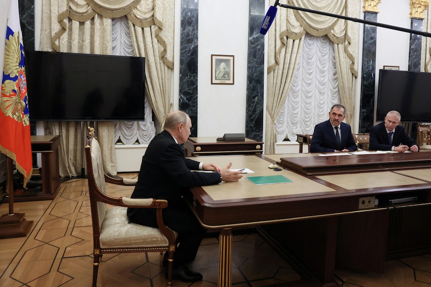 Three men sit at a long table in a decorated conference table