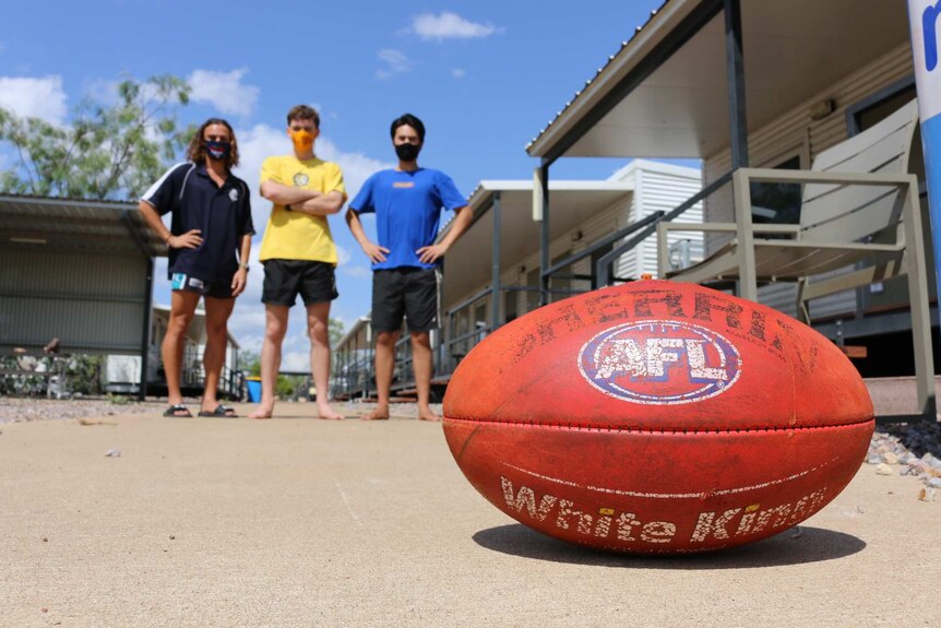 Three boys stand in a sunny outside space with an AFL ball in the foreground.