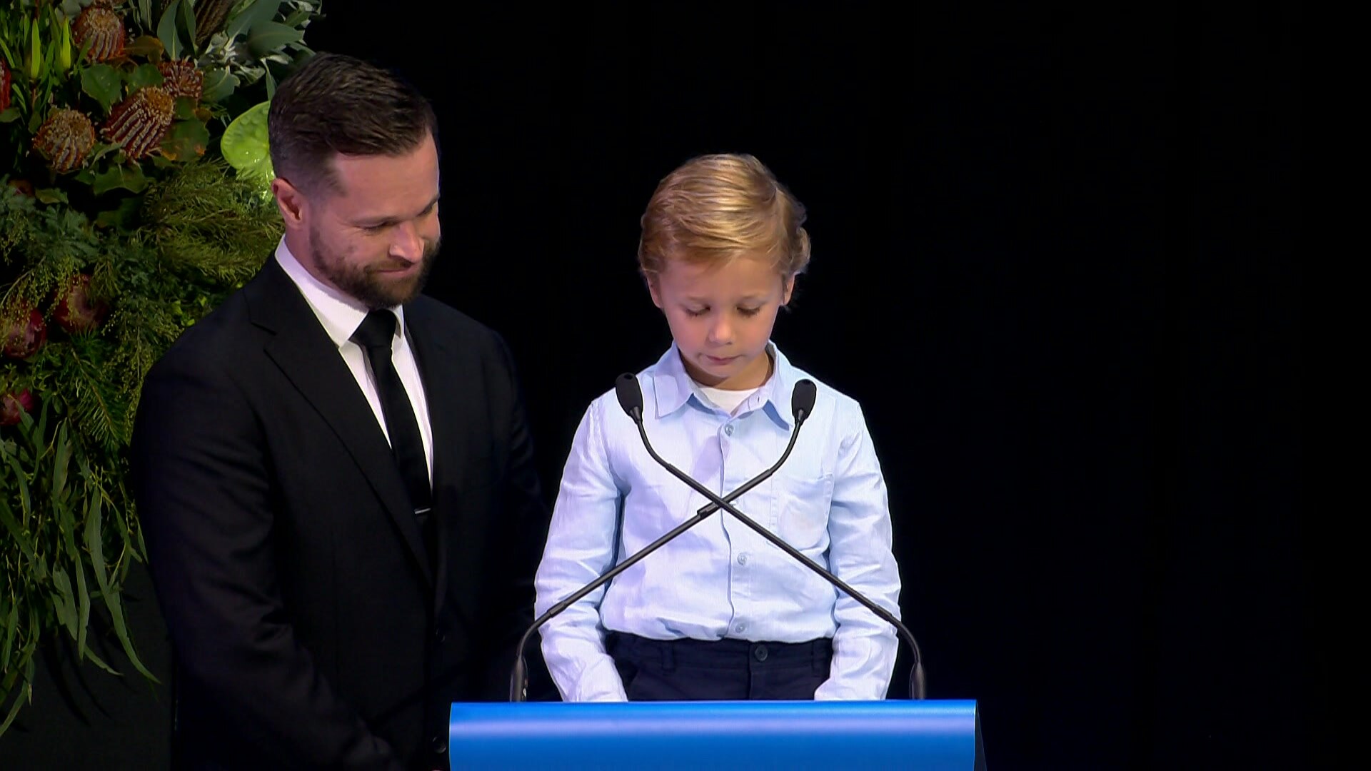 A dark haired man in a black suit stands with a young blond boy in a blue collared shirt who is reading into microphones.