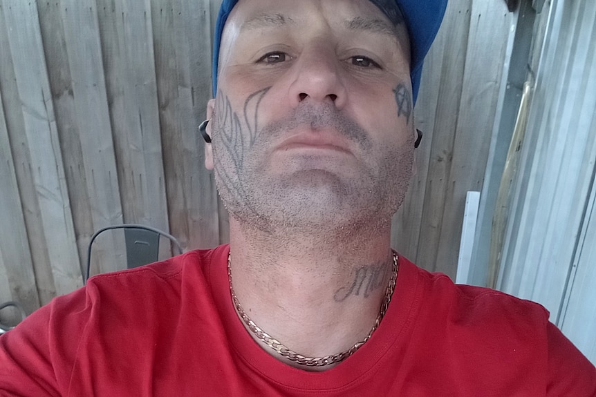 A man with tattoos wearing a red t-shirt and a baseball cap stares at the camera.