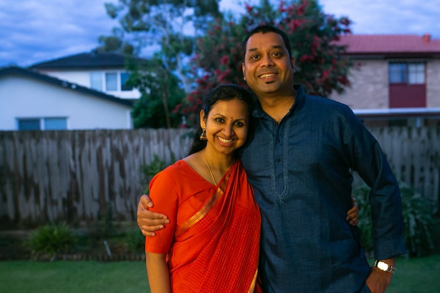 Couple standing in their family home, woman wearing a Sari, man in a blue shirt.
