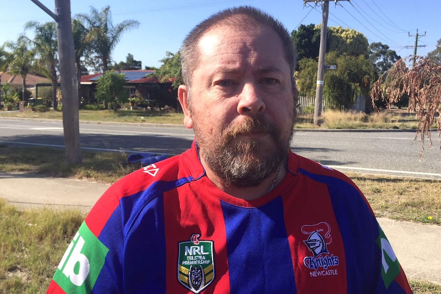 A man in a Newcastle Knights top stands on a street verge, with trees, houses, and powerlines in the background.