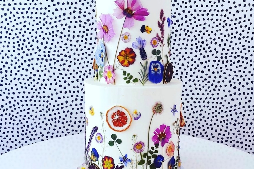 Two tier white frosted cake against a speckled background and decorated with colourful fresh pressed flowers and dried citrus