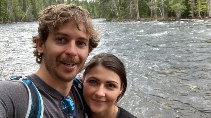 A couple smile at the camera in front of a river