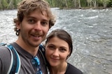 A couple smile at the camera in front of a river