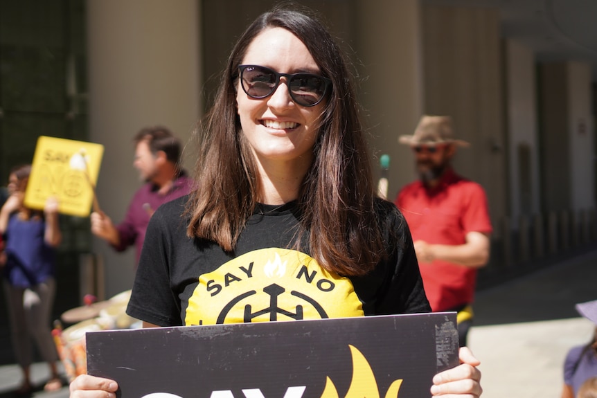 Woodside protest organiser Rhiannon Hardwicke smiles posing for a photo wearing sunglasses and holding a sign.