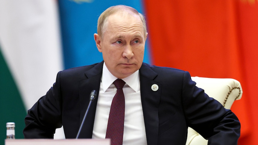 Vladimir Putin, wearing a dark suit, looks ahead as he sits in front of green, white, blue and red banners.