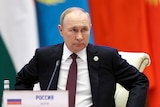 Vladimir Putin, wearing a dark suit, looks ahead as he sits in front of green, white, blue and red banners.