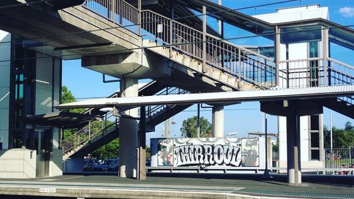 Stairs and lifts at a train station with a graffiti-style sign on a wall in the background that reads 'Thirroul'.