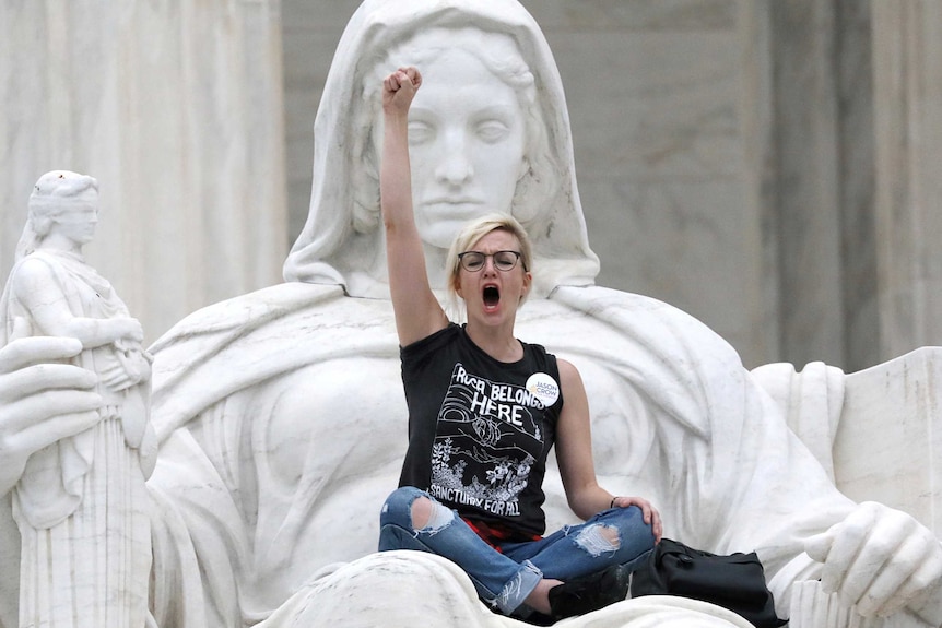 A woman with one fist raised in the air wearing glasses, a black tshirt and jeans, sitting on a white marble statue