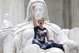 A woman with one fist raised in the air wearing glasses, a black tshirt and jeans, sitting on a white marble statue