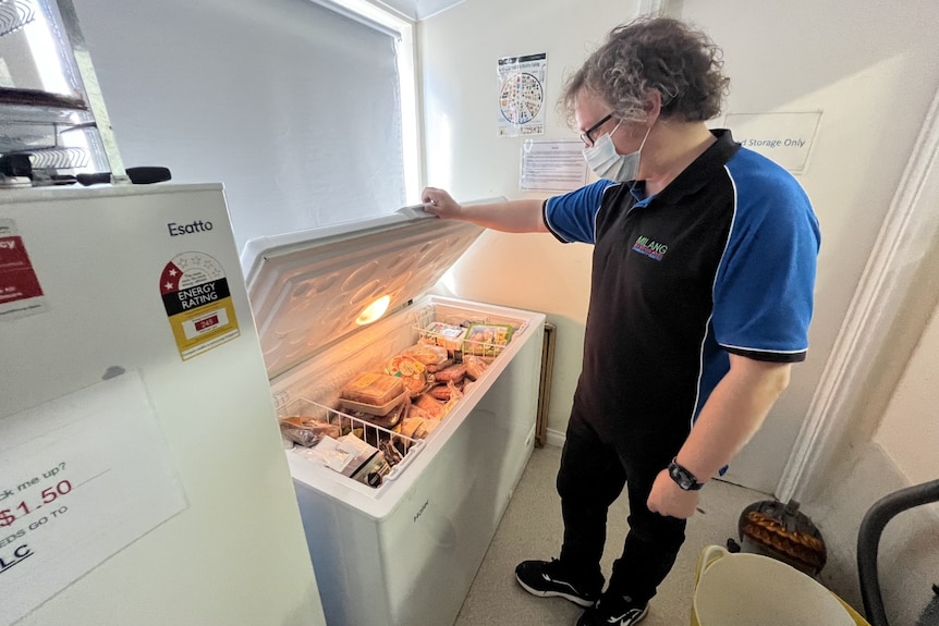 A man wearing a mask, jeans and a polo shirt holds a large freezer packed with food open