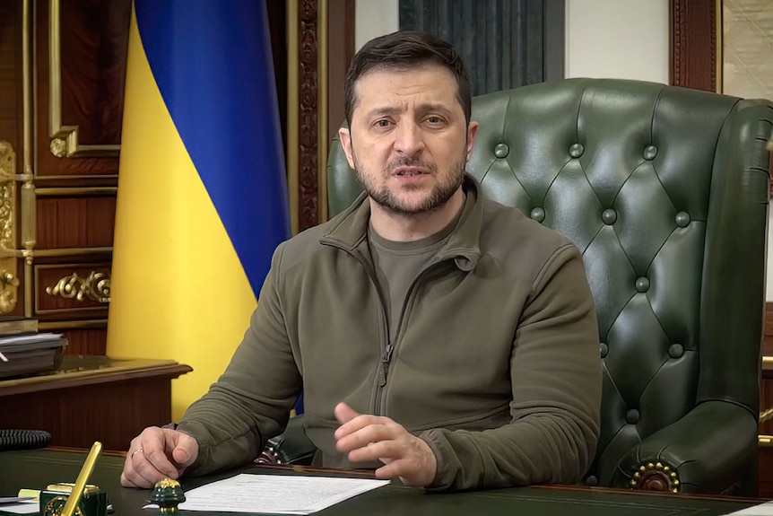 A middle-aged man with beard and khaki clothing sits in green chair in office with Ukraine flag behind him and talks to camera