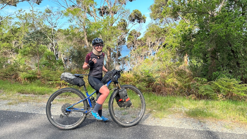 A woman smiles at the camera. She sits on her bicycle. In the background Australian bush with water