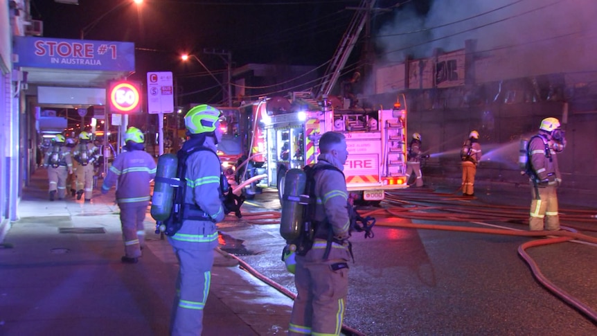 A large group of firefighters stand in the street. There is a fire truck and they're hosing water on the building.