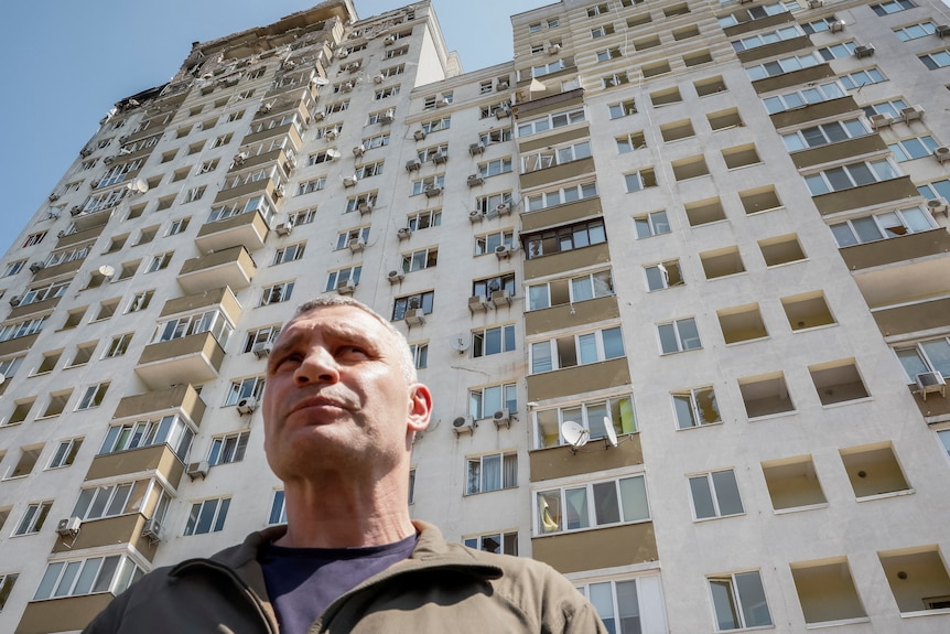 Vitali Klitschko stnds in front of a damaged apartment building