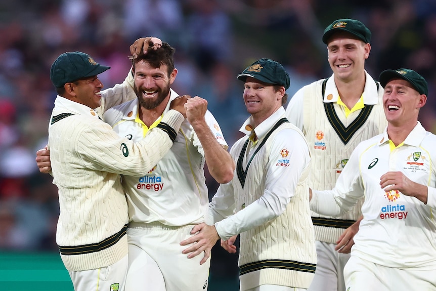 Australian cricketers hug and pat a smiling bowler on the head as everyone celebrates a Test wicket.
