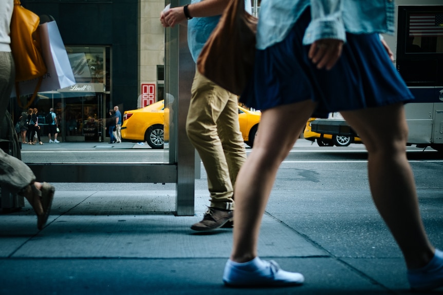 From waist-level down, people in pants and skirts walk along a street with cars driving behind them.