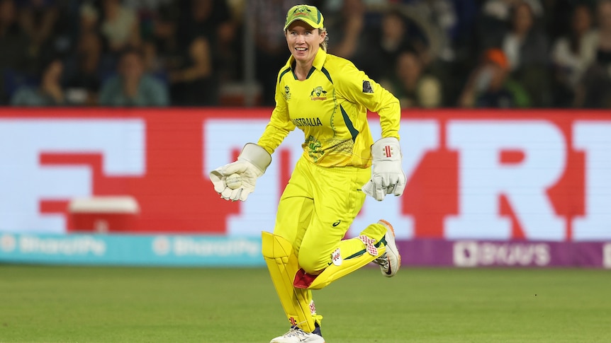 A smiling Australian wicketkeeper, Beth Mooney, runs in holding a ball in her glove after a dismissal.