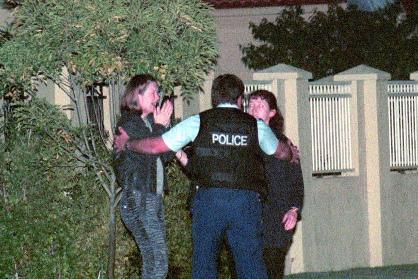 Gennadi Bernovski's wife holds her hands up to her face, with her mouth open, as a police officer puts his hand on her shoulder.