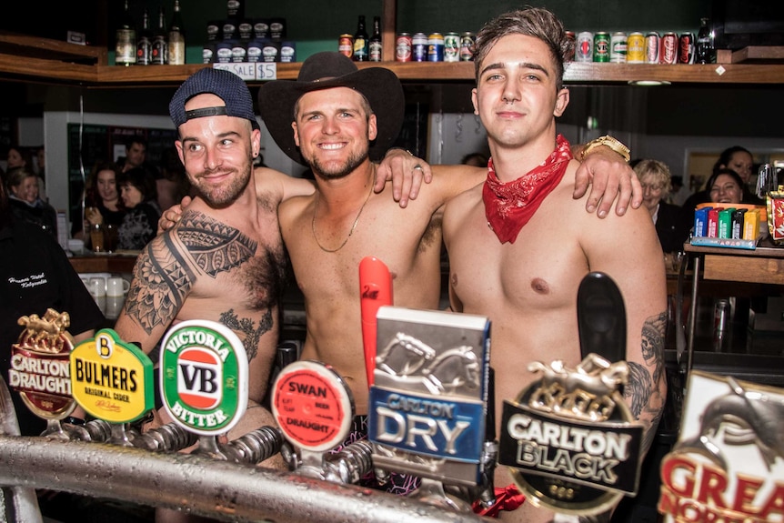 Three shirtless male bartenders smile behind the bar.