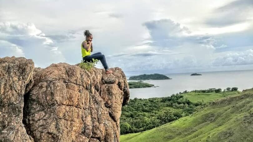This image show the rolling hills in PNG's Port Moresby with a woman sitting atop of cliff look out to the ocean.