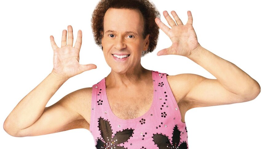 Richard Simmons smiles at the camera with hands next to his face in a pink singlet