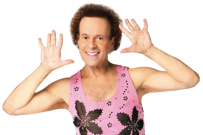 Richard Simmons smiles at the camera with hands next to his face in a pink singlet