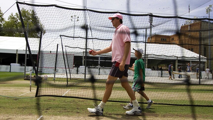 A tall man carrying a cricket ball walks with a young boy by his side an out-of-focus net is seen in the foreground
