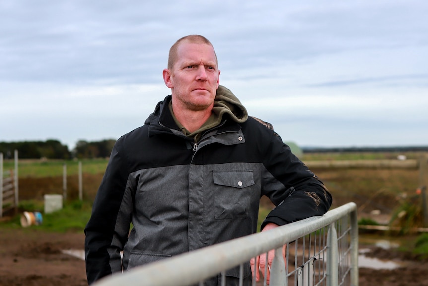 Man in jacket stands resting on fence with farm behind him