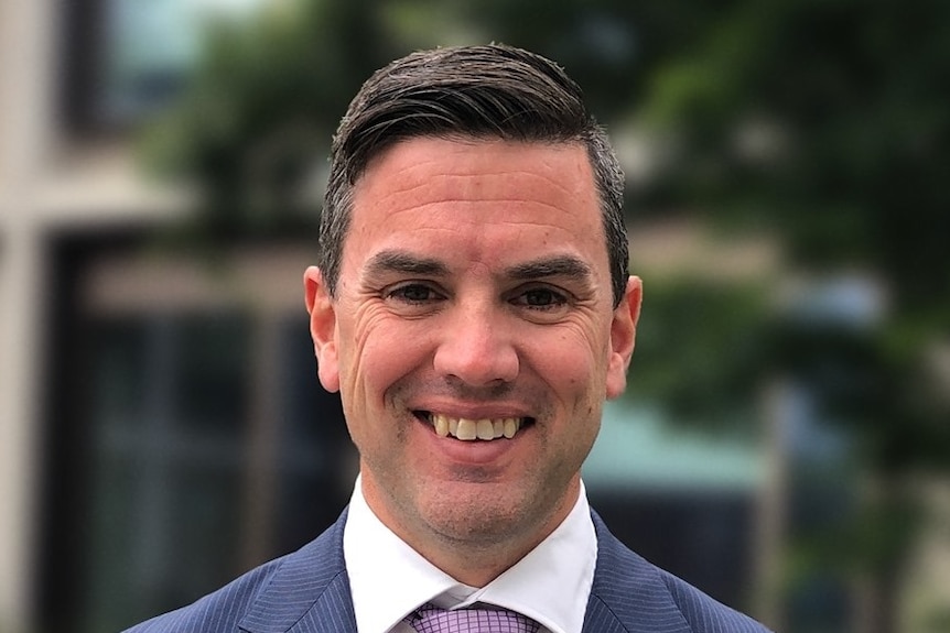 A man in a blue suit with dark hair poses for a photo while smiling.