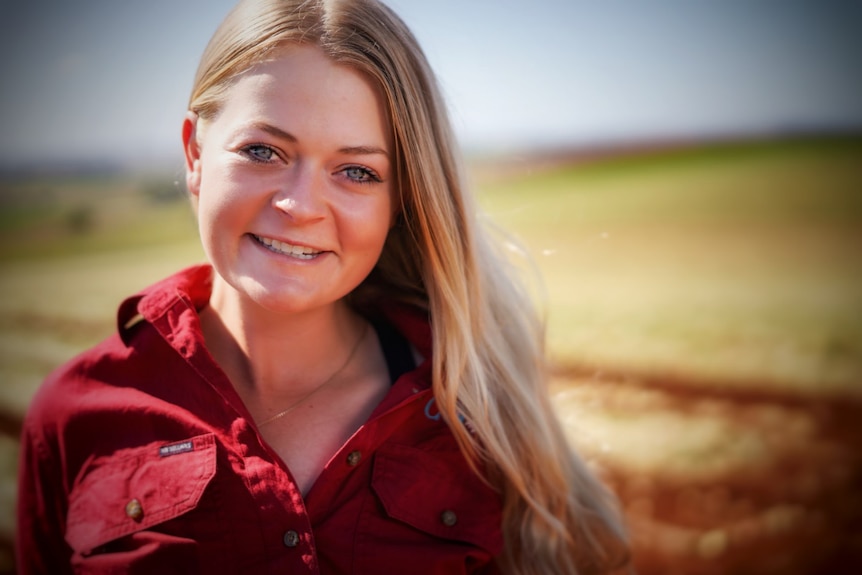 A young blonde woman wearing a red work shirt stands outside, smiling.