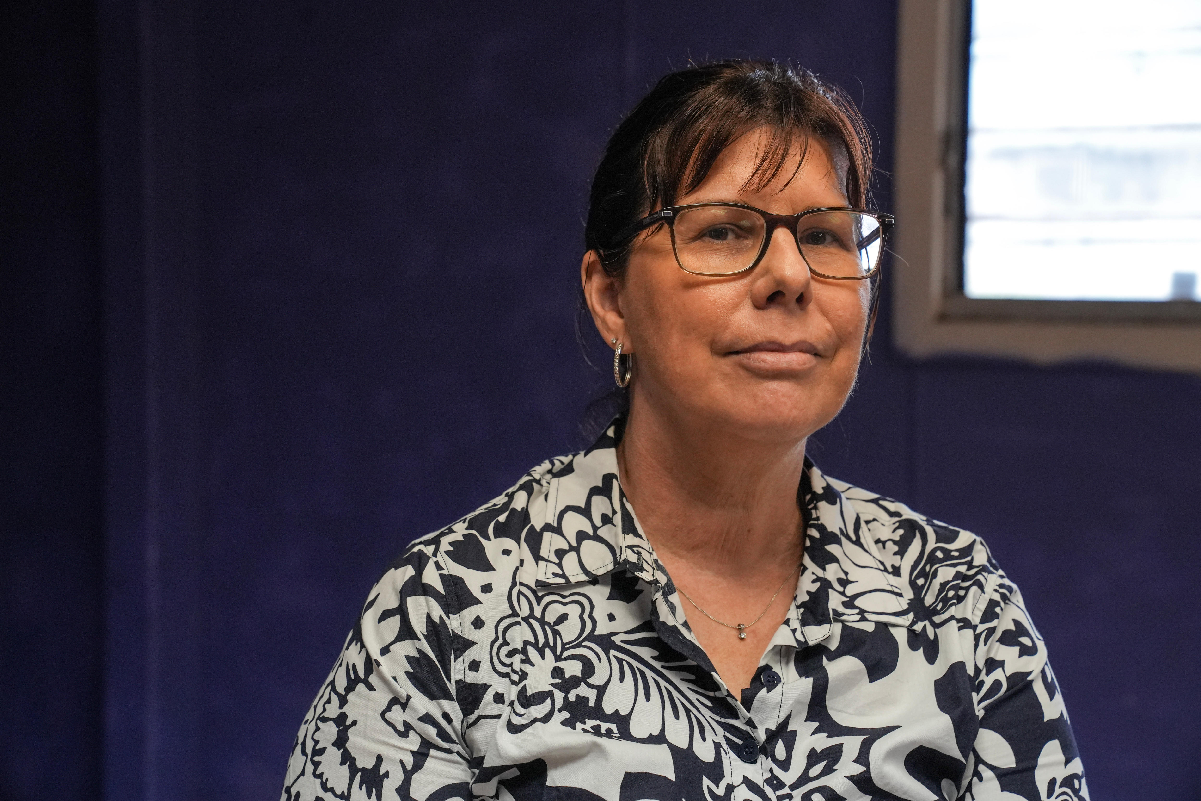 一位戴着眼镜、棕色头发、穿着黑白花纹衬衫的女子，神情严肃地看着镜头。