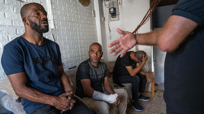 A man in a blue t-shirt looking stunned while sitting with two other men on a couch