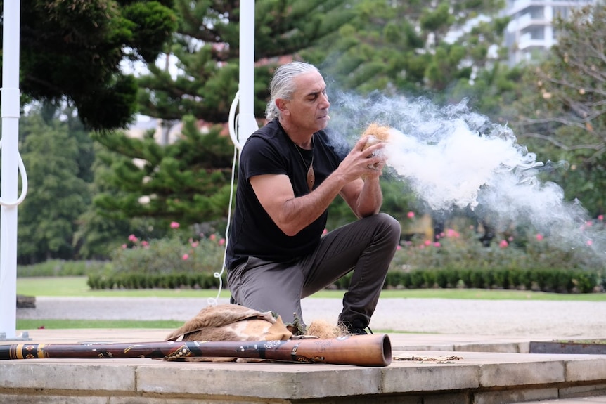 An indigenous man kneels on the ground blowing smoke