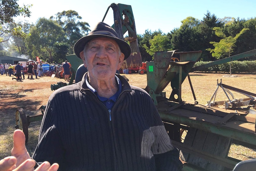 A man in a hat stands in front of an old tractor plough