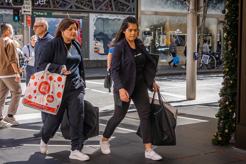 Two women, both with brown hair and wearing black, walk in the city holding shopping bags.