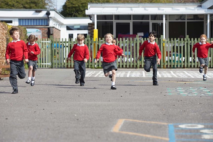 Six primary school students in uniform run towards the camera across a playground