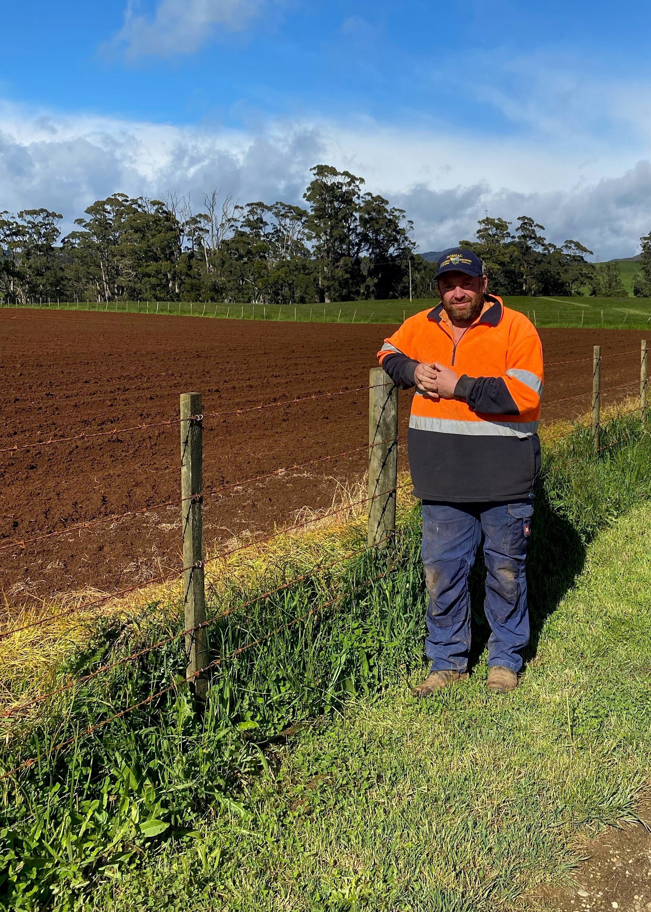 A man in a high visibility jumper stands by a plowed field of red earth. 