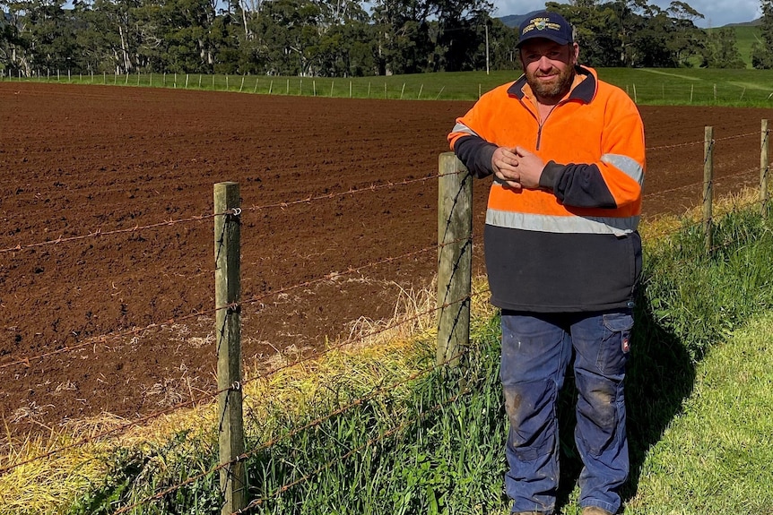 A man in a high visibility jumper stands by a plowed field of red earth. 