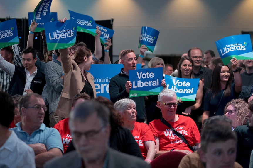 A crowd of people wearing blue hold signs supporting the Liberal Party. Labor supporters in red clothes sit in front of them.  