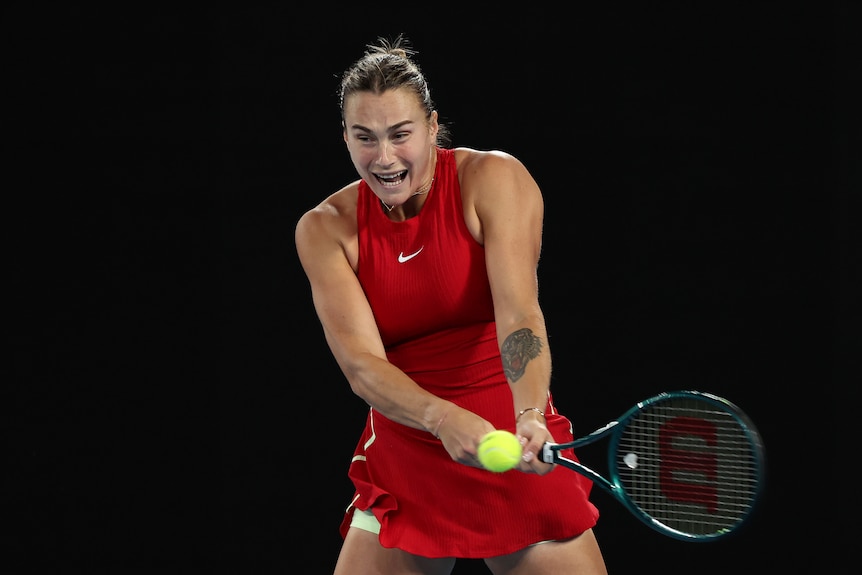 Aryna Sabalenka, in a red top and sport skirt, hits a double-handed backhand, during a night match