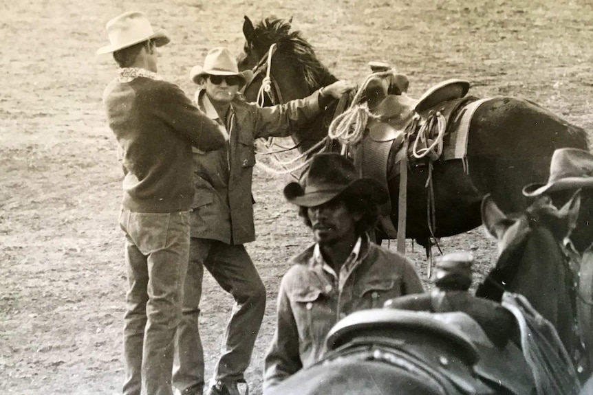 Two men, one with a cigarette, standing on the ground next to a horse in a rodeo arena.