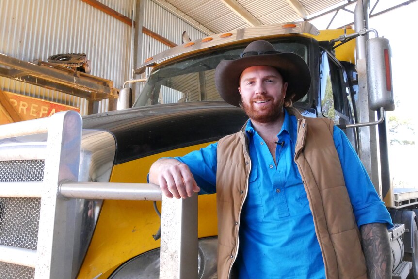 A man in a blue shirt leans against the bull bar of a yellow and black truck.
