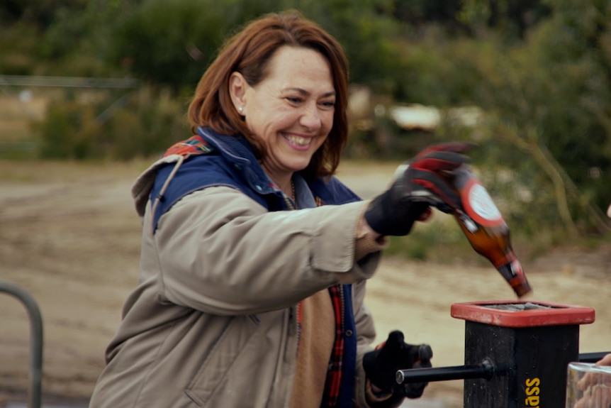 Woman in winter clothing laughing and pushing a glass beer bottle into a metal vertical tube. 