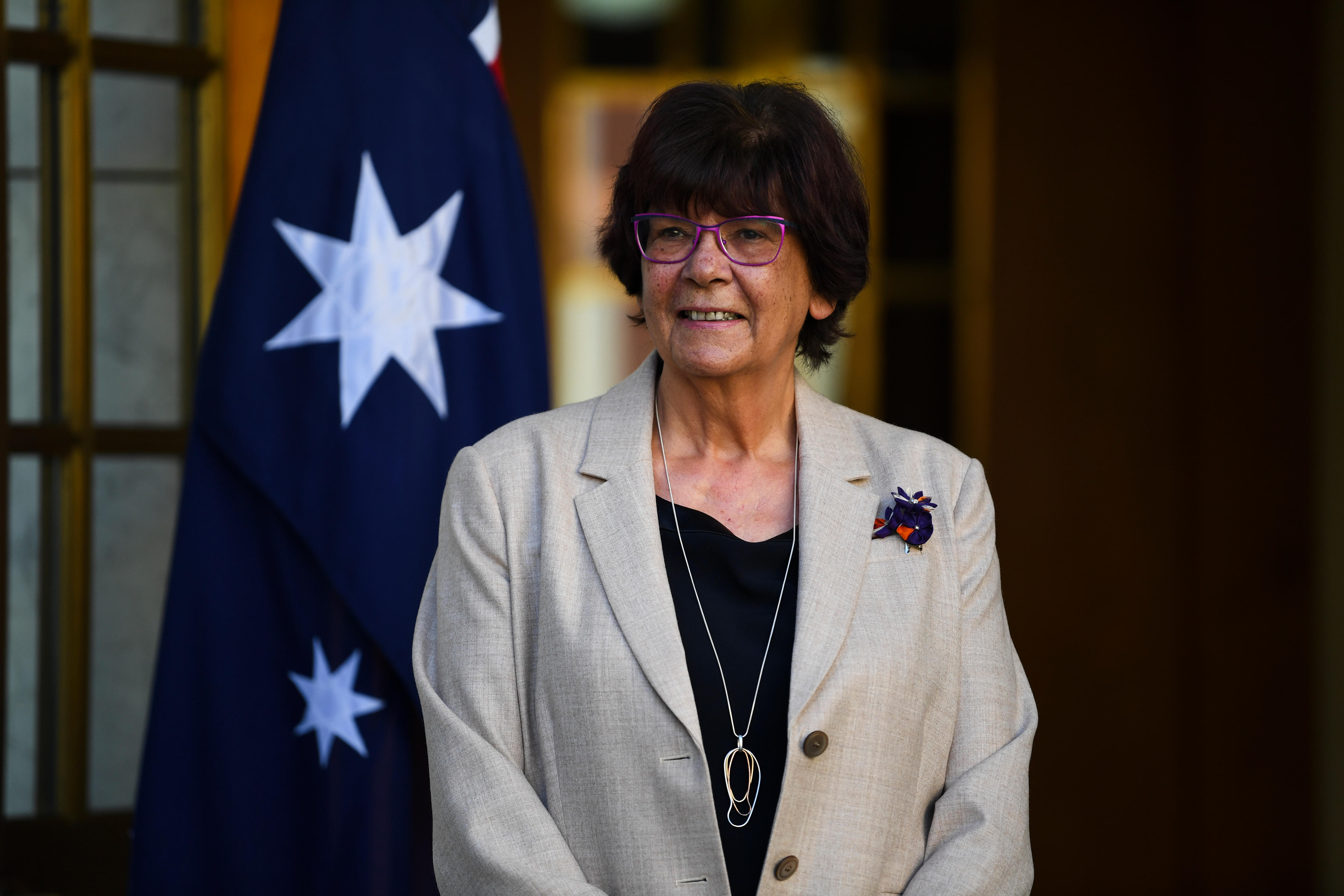 Pat Turner is wearing a beige jacket and standing in front of an Australian flag