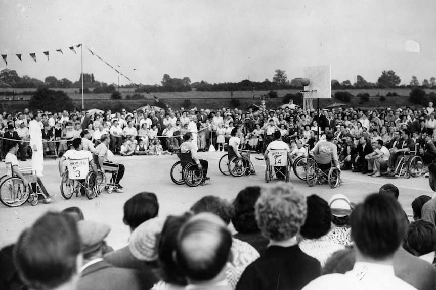 A group of players in wheelchairs play basketball in the open while a crowd looks on.