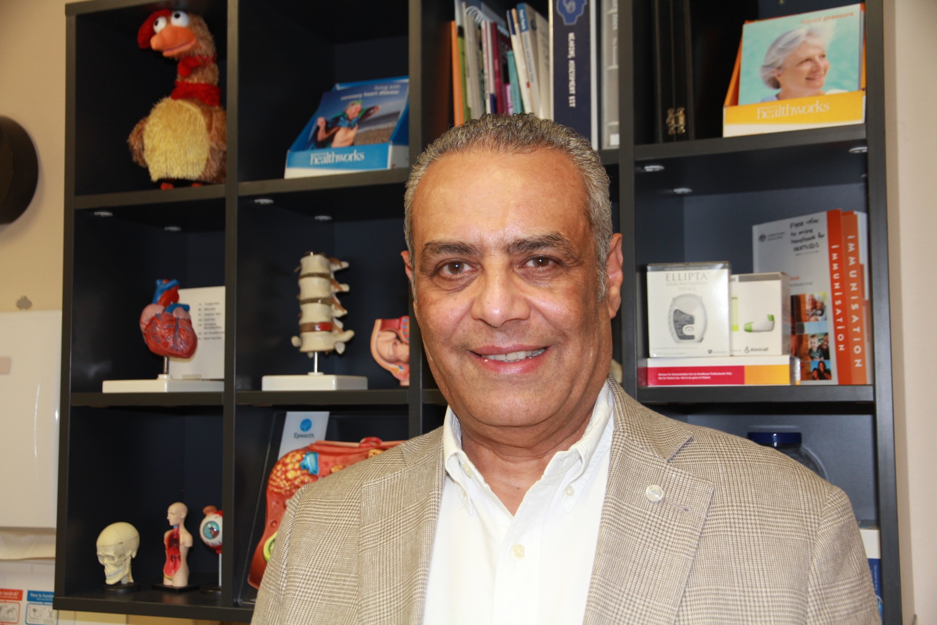 Homem de terno biege em frente a uma prateleira cheia de parafernália médica em cima