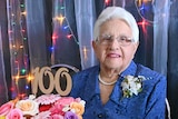 A lady smiles, sitting behind her 100th birthday cake.